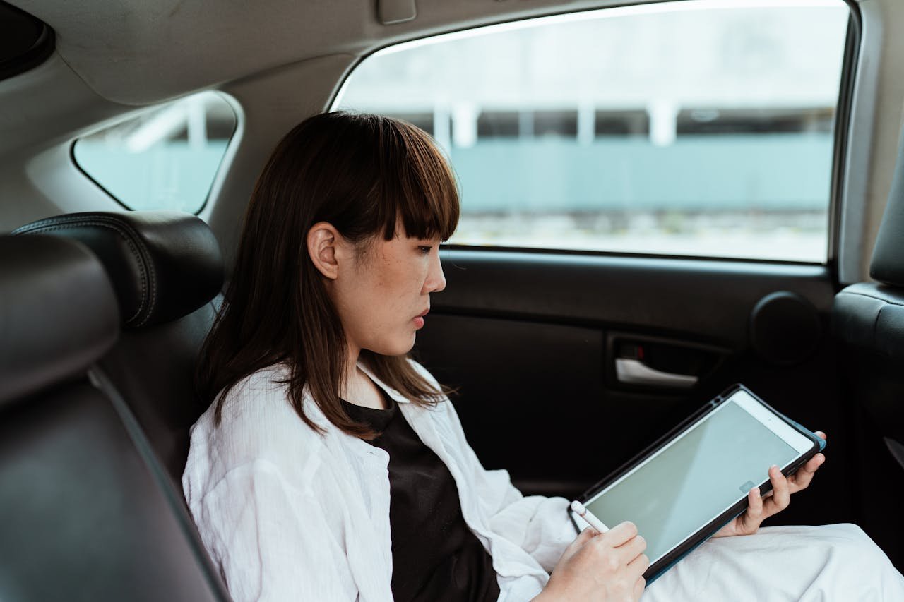 Side view serious Asian female in casual outfit using tablet with empty screen and stylus while riding in car backseat on daytime